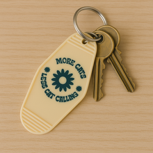 Load image into Gallery viewer, More Cats, Less Cat Calling Keychain
Equal parts protest and purr.

A retro motel-style tag with zero tolerance for street sleaze. Molded in buttercream plastic with a punch of deep green text and a bold daisy, it says what you’re thinking: MORE CATS, LESS CAT CALLING.

Clip it to your keys like brass knuckles for the modern feminist.  Fierce, unapologetic, loud, feral and decidedly not here for your unsolicited commentary.
	•	3.8” x 2” vintage motel shape
	•	Chunky molded plastic with raised lettering
	•	D